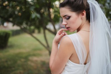 Şık gelinliğini, zarif duvağını ve kusursuz makyajını sergileyen göz kamaştırıcı yakın plan bir gelin. Yeşilliğin yumuşak arkaplanı huzurlu ve romantik bir ortam yaratır..