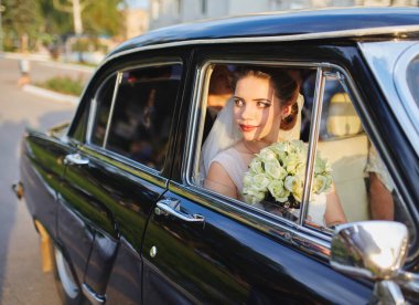 Siyah bir arabanın içinde oturan gelin elinde bir buket beyaz gülle pencereden dışarı dikkatlice bakıyor. Gün batımında yakalanmış, sakin ve zarif gelinliğini vurguluyor..