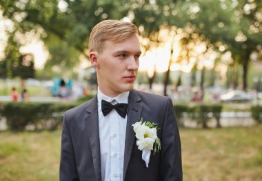 Klasik siyah takım elbiseli ve papyonlu damadın portresi. Beyaz çiçekli yaka çiçeğiyle tamamlanmış. Yeşil ve yumuşak günbatımının bulanık arka planında duruyor..