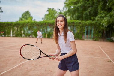 Genç bir bayan tenisçi raketini güvenli bir şekilde tutarken, takım arkadaşı arka planda hazırlanıyor. Yeşillik, açık hava atmosferini güçlendiriyor..