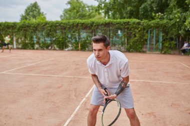 Odaklanmış genç bir adam kil tenis kortunda servis yapmaya hazırlanıyor. Beyaz polo tişört ve gri şort giymiş, elinde bir tenis raketi tutmakta ve harekete geçmeye hazırdır..