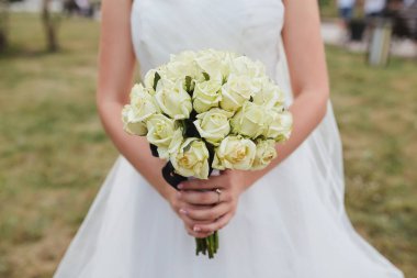 Çiçeklere ve zarif gelinliğine odaklanmış, açık hava arka planına sahip bir buket beyaz gül tutan bir gelinin yakın çekimi..