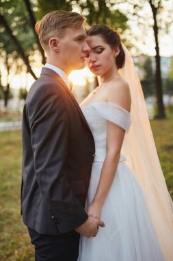 Yeni evliler gün batımında özel bir anı paylaşıyorlar. Gelin beyaz bir elbise ve duvak içinde bir buket beyaz gül tutarken damat nazikçe onu yanında tutuyor..