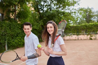Bir erkek ve bir kadın tenis kortunun önünde kameraya güvenle gülümsüyor. Takım çalışması ve dostluk yayarlar, eğlenceli bir maça ya da antrenmana hazırdırlar..