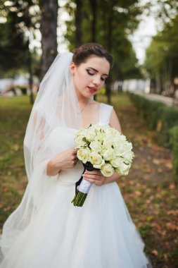 Çiçeklere ve zarif gelinliğine odaklanmış, açık hava arka planına sahip bir buket beyaz gül tutan bir gelinin yakın çekimi..