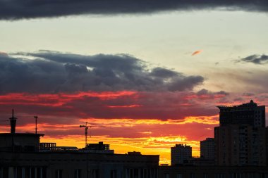 Yoğun turuncu ve sarı renkli canlı bir günbatımı, binaların siluetlerini ve bir inşaat vincini ön plana çıkarıyor. Bulutlu bir gökyüzünün altında dramatik bir ufuk çizgisi oluşturuyor..