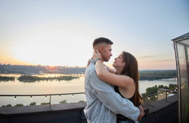 Romantik bir çatı randevusu sırasında genç bir çift öpüşür ve kucaklaşır. Gün batımında şehrin arka planında..