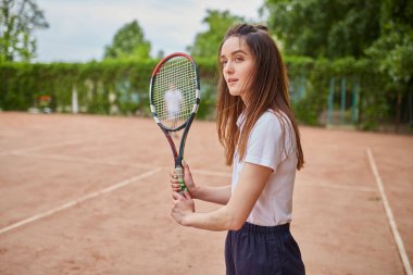 Genç bir bayan tenisçi kil sahada kararlılıkla hazırlanıyor. Beyaz polo ve donanma şortu giymiş, raketini sıkı sıkı tutuyor, yemyeşil bir ortamda harekete geçmeye hazır..