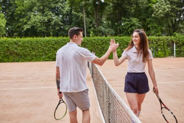 Bir erkek ve kadın kil tenis kortunda beşlik çakarak takım çalışmalarını kutluyor ya da başarılarını paylaşıyorlar. İkisi de gülümsüyor, raket tutuyor ve açık hava ortamının tadını çıkarıyor..
