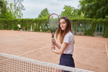 Genç bir bayan tenisçi kil sahada kararlılıkla hazırlanıyor. Beyaz polo ve donanma şortu giymiş, raketini sıkı sıkı tutuyor, yemyeşil bir ortamda harekete geçmeye hazır..