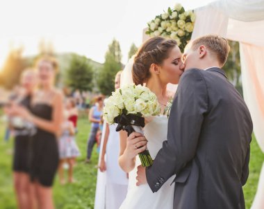Yeni evliler düğün törenlerinde romantik bir öpücük paylaşıyorlar. Gelin, misafirlerle çevrili bir buket beyaz gül ve yumuşak kumaş ve çiçeklerle süslenmiş bir kemer tutuyor..