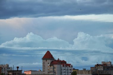 Yükselen bulutlar ve yumuşak akşam ışıklarıyla dolu bir gökyüzünün altında huzurlu bir şehir. Binalar dramatik bulut oluşumlarıyla tezat oluşturarak barışçıl ama dinamik bir şehir manzarası yaratıyor..