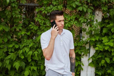 Dışarıda duran genç bir adam kulağına akıllı telefon dayayıp yeşil sarmaşık arka planında beyaz polo tişört giyiyor. Sahne, odak ve sıradan bir atmosfer taşır..