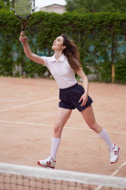 Yoğun bir maç sırasında kadın bir tenisçi kil sahada forehand salıncak yapıyor. Dinamik hareket ve odaklanma onun yaz sporlarındaki tutkusunu ve atletizmini vurguluyor..