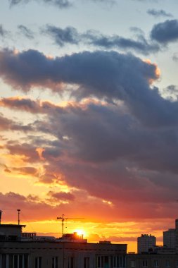 Dramatik bir turuncu ve kırmızı gün batımına karşı inşa edilmiş bir vinç silueti. Güneş ufkun altına batarken, katmanlı bulutlarla dolu canlı gökyüzü şehir sahnesine yoğunluk katıyor..