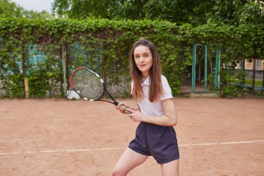 Genç bir kadın kil tenis kortunda hizmet etmeye hazırlanıyor. Elinde beyaz polo ve donanma şortu giymiş bir tenis topu ve raket tutuyor..