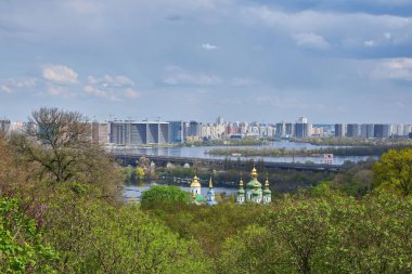 Kyiv 'in çarpıcı manzarası, tarihi bir kilisenin altın kubbelerini sergiliyor, yemyeşil ağaçlarla çevrili, modern şehir silüeti ve arka planda Dnipro Nehri ile..