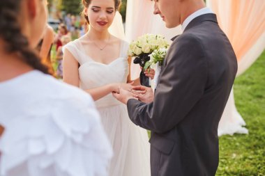Damat tören sırasında gelinin parmağına bir alyans takar. Gelin elinde bir buket beyaz gül tutarken ikisi de süslü bir kemerin altında akan kumaşla yemin ediyor..