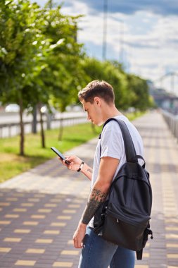 Beyaz tişörtlü ve sırt çantalı genç bir adam nehir kenarındaki korkulukların yanında duruyor, akıllı telefonuna odaklanmış. Arka planda sakin bir nehir, ağaçlar ve bulutlu bir gökyüzü var..