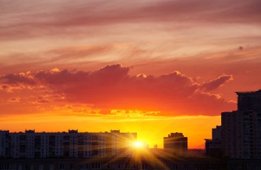 Şehrin ufuk çizgisinde gün batımı. Güneş görünür, bulutların üzerine sıcak bir parıltı saçıyor. Binalar renkli gökyüzüne karşı siluetlidir..