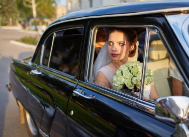 Beyaz bir duvak takan, şık bir elbise giyen, klasik siyah bir arabanın içinde oturan güzel bir gelin. Güneş ışığı, pencereden bakarken onun huzurlu ve neşeli yüzünü hafifçe aydınlatıyor..