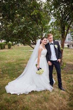 Gelin beyaz bir gül buketi tutuyor ve damat onu kucaklıyor, ceketini omuzlarının üzerine örtüyor. Altın güneş ışığıyla serin bir park arkaplanı romantik bir ortama ekliyor..