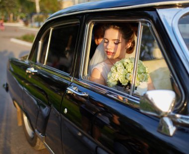 Beyaz bir duvak takan, şık bir elbise giyen, klasik siyah bir arabanın içinde oturan güzel bir gelin. Güneş ışığı, pencereden bakarken onun huzurlu ve neşeli yüzünü hafifçe aydınlatıyor..
