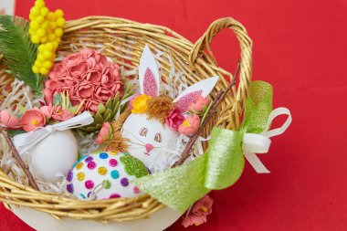 Süslü Paskalya yumurtaları, yapay çiçekler ve kırmızı bir yüzey üzerinde duran dekoratif elementlerle dolu bir hasır sepet..
