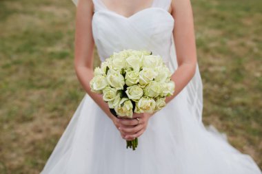 Çiçeklere ve zarif gelinliğine odaklanmış, açık hava arka planına sahip bir buket beyaz gül tutan bir gelinin yakın çekimi..