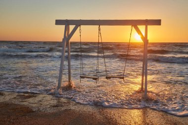 Kumlu kumsalda beyaz ahşap bir salıncak canlı bir günbatımında güçlü okyanus dalgalarıyla yüzleşir..