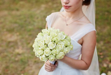 Beyaz gelinlik içinde elinde bir buket beyaz gül tutan bir gelin. Çekim çiçeklere ve elbisenin üst kısmına odaklanır, bulanık çimen arka planıyla..
