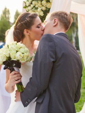 Yeni evliler düğün törenlerinde romantik bir öpücük paylaşıyorlar. Gelin, misafirlerle çevrili bir buket beyaz gül ve yumuşak kumaş ve çiçeklerle süslenmiş bir kemer tutuyor..