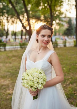 Güzel bir gelin elinde bir buket beyaz gül tutarak gülümser. Arka planda ağaçların arasından süzülen güneş ışığı açık havada sıcak ve romantik bir düğün sahnesi yaratır..