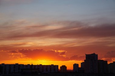Şehrin ufuk çizgisinde gün batımı. Güneş görünür, bulutların üzerine sıcak bir parıltı saçıyor. Binalar renkli gökyüzüne karşı siluetlidir..