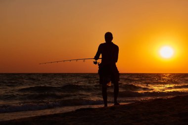 Sahilde duran bir balıkçının silueti güneş denizin üzerinde batarken bir olta atıyor..