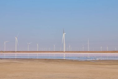 Açık bir gökyüzünün altında, ufukta rüzgar türbinleri olan canlı pembe bir tuz gölü..