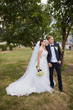 Gelin beyaz bir gül buketi tutuyor ve damat onu kucaklıyor, ceketini omuzlarının üzerine örtüyor. Altın güneş ışığıyla serin bir park arkaplanı romantik bir ortama ekliyor..