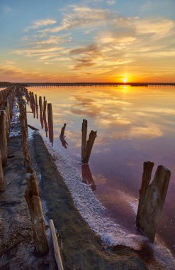 Sakin, pembe bir tuz gölünün üzerinde canlı bir günbatımı. Suya yansıyan ahşap yapıların kalıntıları..