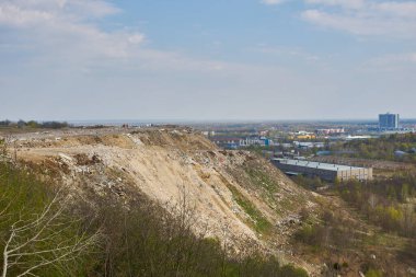 Arkasında büyük bir enkaz yığını, toprak yollar ve sanayi binaları olan bir inşaat atığı manzarası.