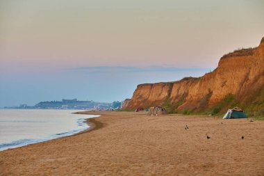 Kıyı kayalıklarının yakınındaki kumlu bir plajda kamp çadırları kurulmuş. Deniz üzerinde canlı bir gündoğumu varmış..
