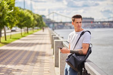 Beyaz tişörtlü ve kot pantolonlu genç bir adam, siyah bir sırt çantası taşıyor, nehir kenarındaki bir patikada korkuluklara yaslanıyor. Arka planda ağaçlar ve bir köprü görünür..