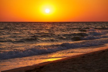 Dalgalar okyanusun üzerinde canlı kırmızı bir günbatımının altında kıyıya doğru yuvarlanıyor. Doğal, düzenlenmemiş fotoğraf..