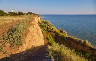 Sahildeki kayalıkların tepesinden denize bakan bir set merdiven, uzaktan görünen bir deniz feneri. İçerik başına Cloud Grass içerir, ancak belirgin değildir.