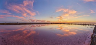 Sakin, pembe bir tuz gölünün üzerinde canlı bir günbatımı. Suya yansıyan ahşap yapıların kalıntıları..