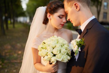 Gelin ve damat gün batımında romantik bir anı paylaşıyorlar. Resmi takım elbiseli damat, geline yakın duruyor. Beyaz bir elbise ve akarsu peçesiyle, altın güneş ışığıyla aydınlatılıyor..
