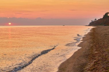 Denizin üzerinde gün doğumu, kumlu bir sahili ve sahil kayalıklarını aydınlatıyor..