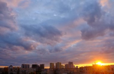 Şehrin ufuk çizgisinde gün batımı. Güneş görünür, bulutların üzerine sıcak bir parıltı saçıyor. Binalar renkli gökyüzüne karşı siluetlidir..