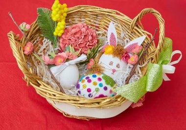 Süslü Paskalya yumurtaları, yapay çiçekler ve kırmızı bir yüzey üzerinde duran dekoratif elementlerle dolu bir hasır sepet..
