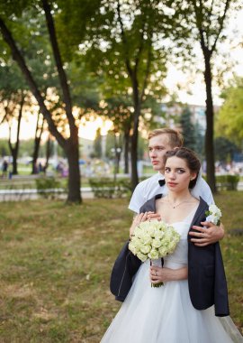 Gelin beyaz bir gül buketi tutuyor ve damat onu kucaklıyor, ceketini omuzlarının üzerine örtüyor. Altın güneş ışığıyla serin bir park arkaplanı romantik bir ortama ekliyor..