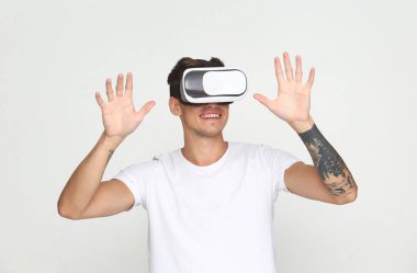 Beyaz tişörtlü genç bir adam VR kulaklık takıyor, bir eliyle öne bakıyor, diğer eliyle kulaklığı tutuyor, sade beyaz bir arka planda izole edilmiş..